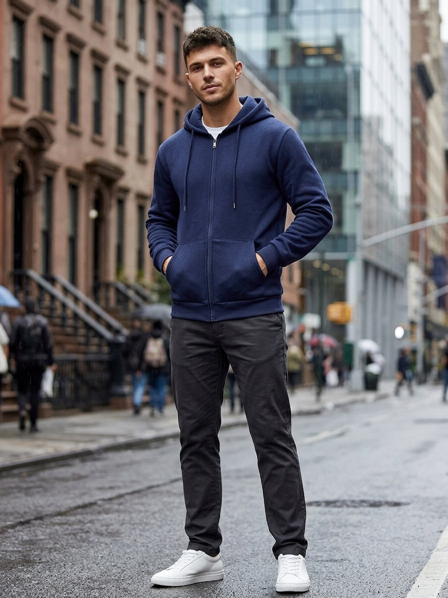 A man standing on a city street wearing a navy blue zip-up hoodie, dark gray pants, and white sneakers.