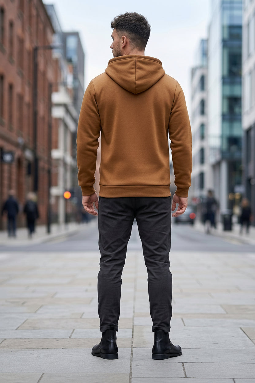 A man viewed from behind wearing a camel brown ribbed hoodie with textured sleeve panels, dark gray trousers, and black boots, standing on a city street.