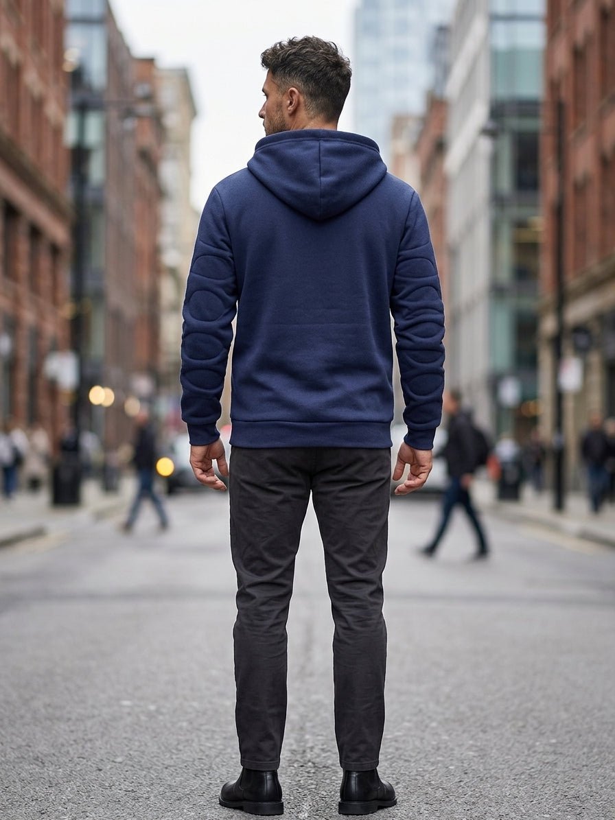 A man viewed from behind wearing a navy blue quilted puffer hoodie, dark gray slim-fit pants, and black boots, standing on a city street with blurred buildings and pedestrians in the background.