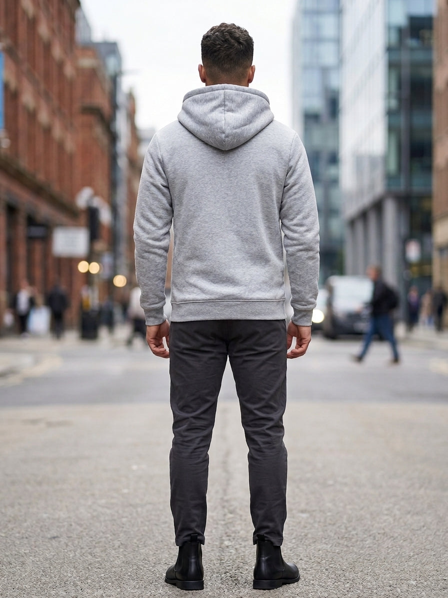 A man viewed from behind wearing a light gray heathered hoodie, dark gray slim-fit trousers, and black boots, standing on a city street.