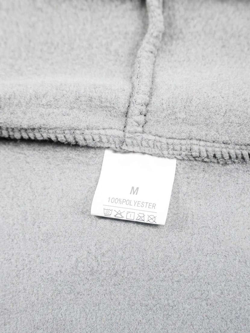 Close-up of a light gray fleece or fabric garment showing a stitched fold and a small white size tag that reads M 100% POLYESTER.