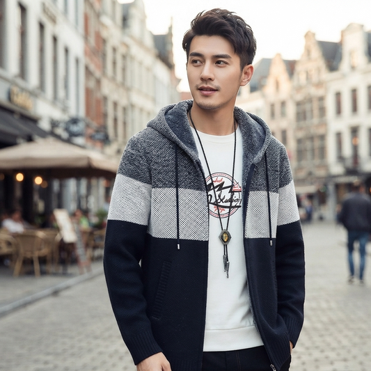 A man wearing a color-blocked knit zip-up hoodie with gray, white, and black stripes, layered over a white crewneck sweatshirt with a graphic print, standing on a cobblestone street.