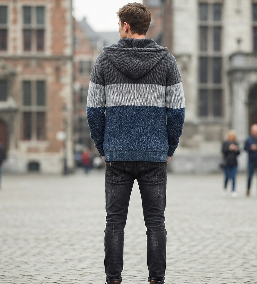 A man standing in a cobblestone town square, viewed from behind, wearing a color-block knit hoodie with dark gray, light gray, and navy blue stripes, paired with dark jeans and brown boots.