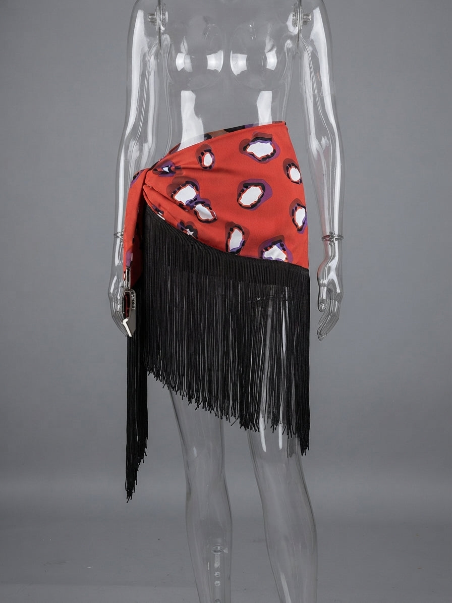 A red wrap-style skirt with black fringe and a black, white, and purple abstract print, displayed on a clear mannequin against a gray background.