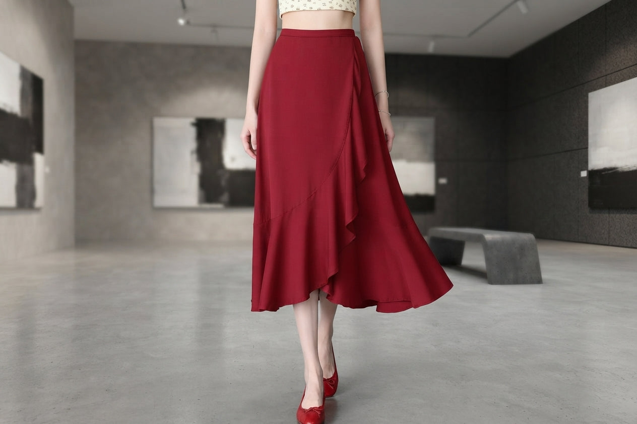 A woman models a high-waisted, mid-length red skirt with an asymmetrical layered ruffle hem, paired with red pointed-toe heels in a modern gallery setting.
