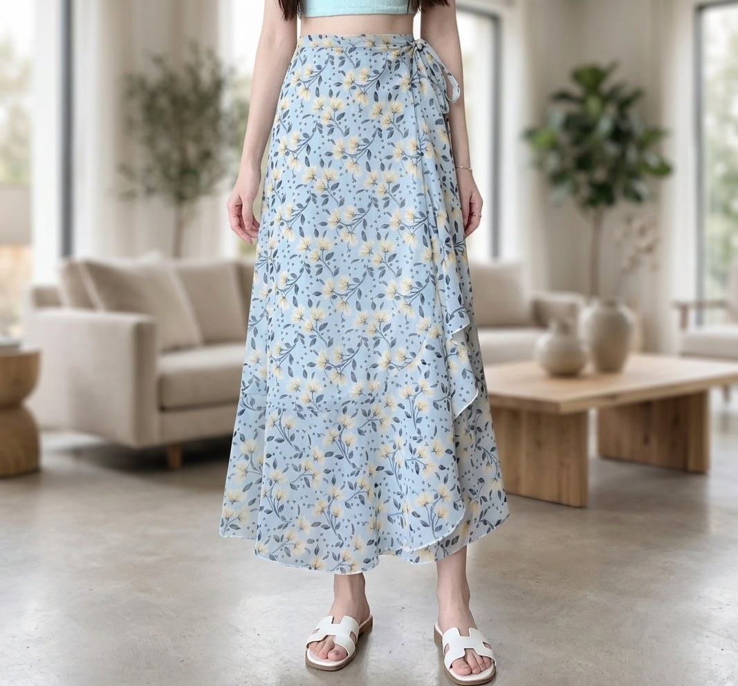 A woman wearing a light blue floral maxi skirt with a tie waist, paired with white slide sandals, standing in a bright, modern living room.