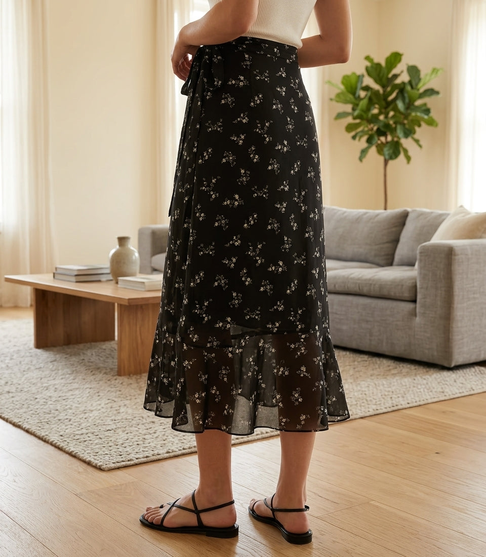 A woman wearing a black midi skirt with a small white floral print, standing in a bright living room with a sofa, coffee table, and potted plant in the background.