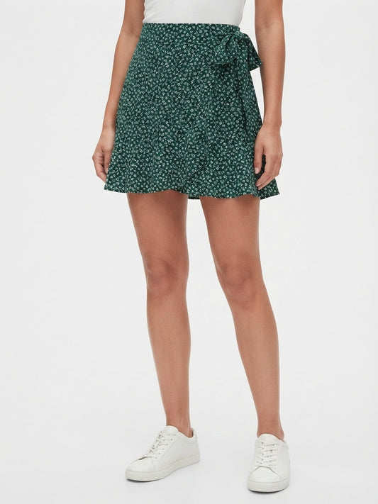 A model wearing a dark green high-waisted mini skirt with a white floral print and a side tie, paired with white sneakers against a plain white background.