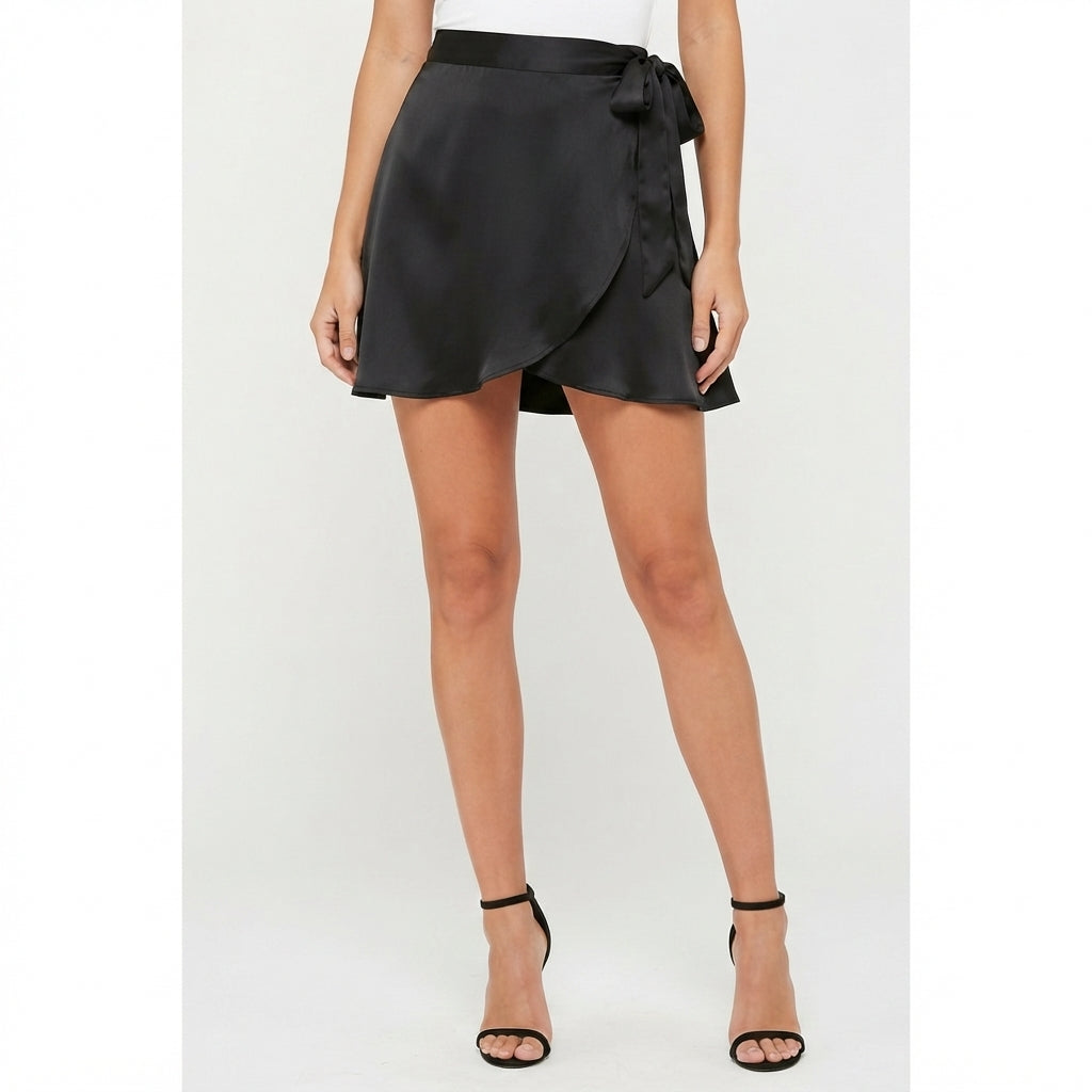 A black satin wrap mini skirt with a matching waistband and side bow, shown on a model wearing black strappy high-heeled sandals against a light gray studio background.