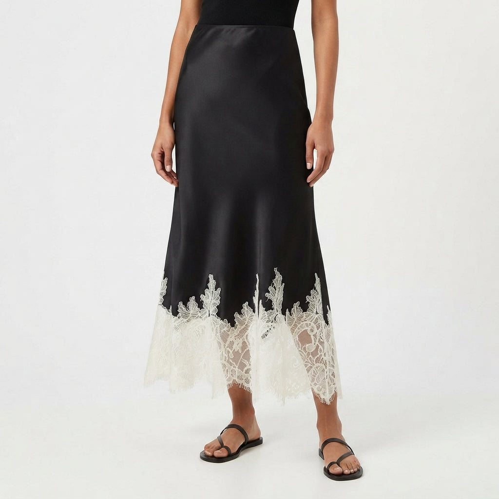 A model wearing a black satin midi skirt with a white lace hem, paired with black flat sandals, against a plain white studio background.