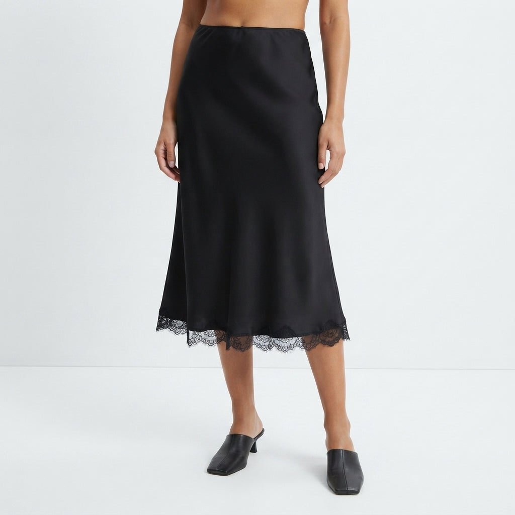 A model wearing a black satin midi skirt with a lace hem, paired with black mule heels, against a clean white studio background.