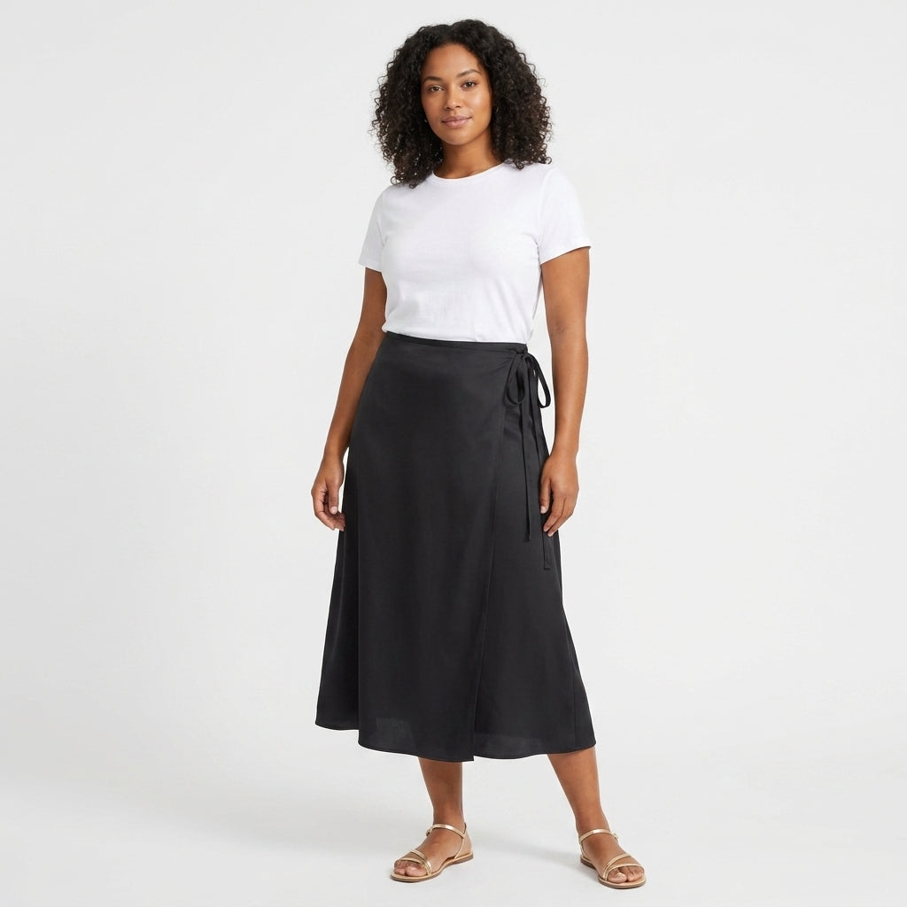 A woman modeling a black wrap midi skirt with a side tie, paired with a white t-shirt and flat sandals, against a plain white studio background.