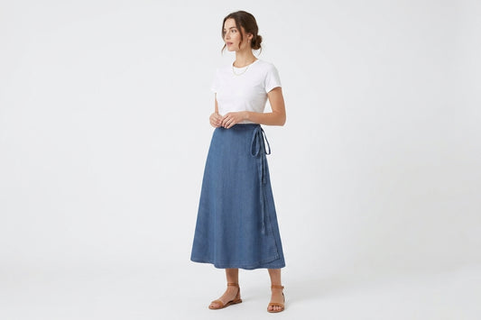 A woman modeling a blue denim midi skirt with a tie waist, paired with a white t-shirt and brown flat sandals, against a plain white studio background.