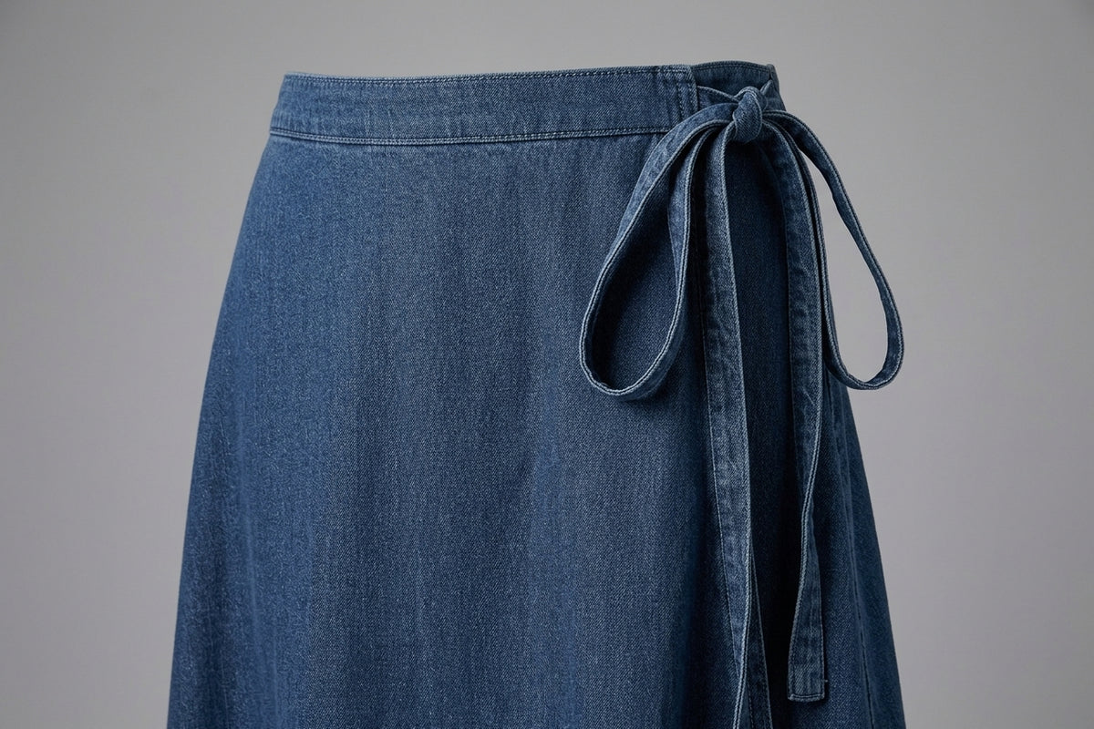 A close-up of a blue denim skirt with a matching fabric tie belt at the waist, shown against a neutral gray background.