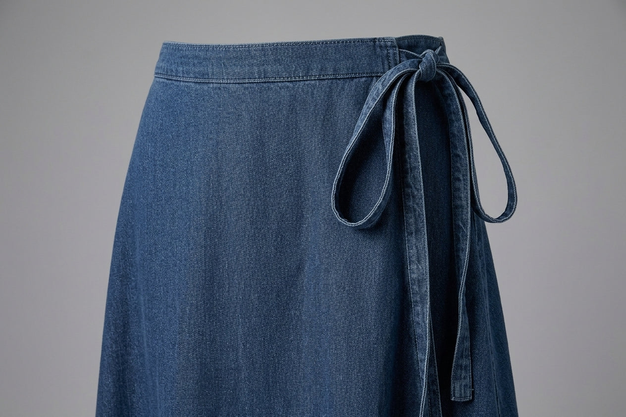 A close-up of a blue denim skirt with a matching fabric tie belt at the waist, shown against a neutral gray background.
