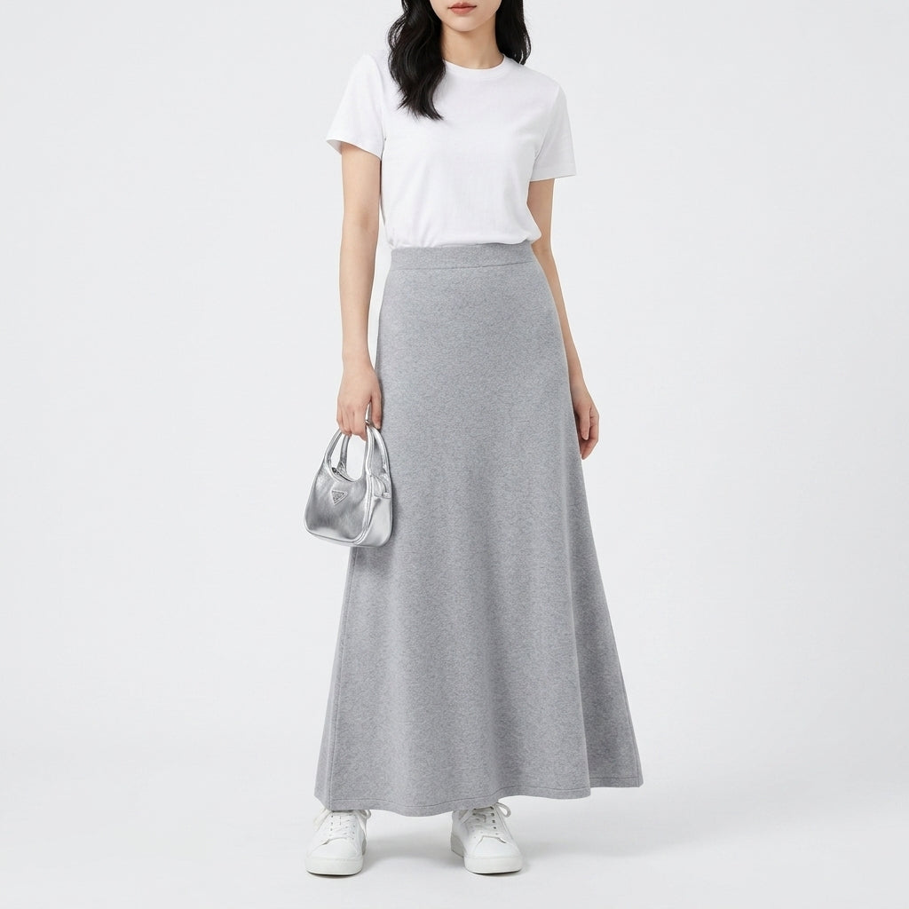 A model wearing a white short-sleeve t-shirt and a high-waisted, flowing light gray maxi skirt, holding a small metallic silver handbag against a plain white studio background.