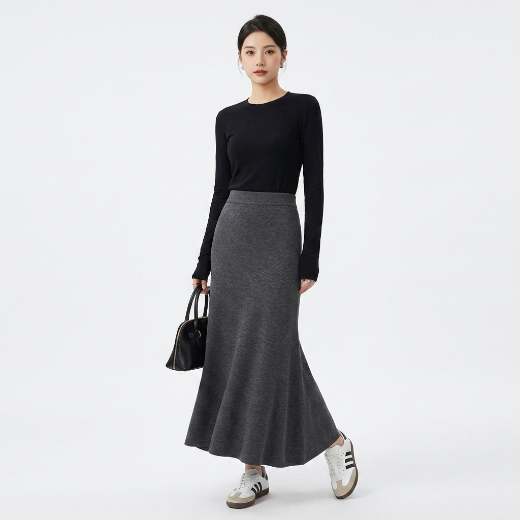 A woman modeling a black long-sleeve crewneck top and a high-waisted, flared dark gray knit midi skirt, holding a small black handbag and wearing white sneakers with black stripes, against a plain white studio background.