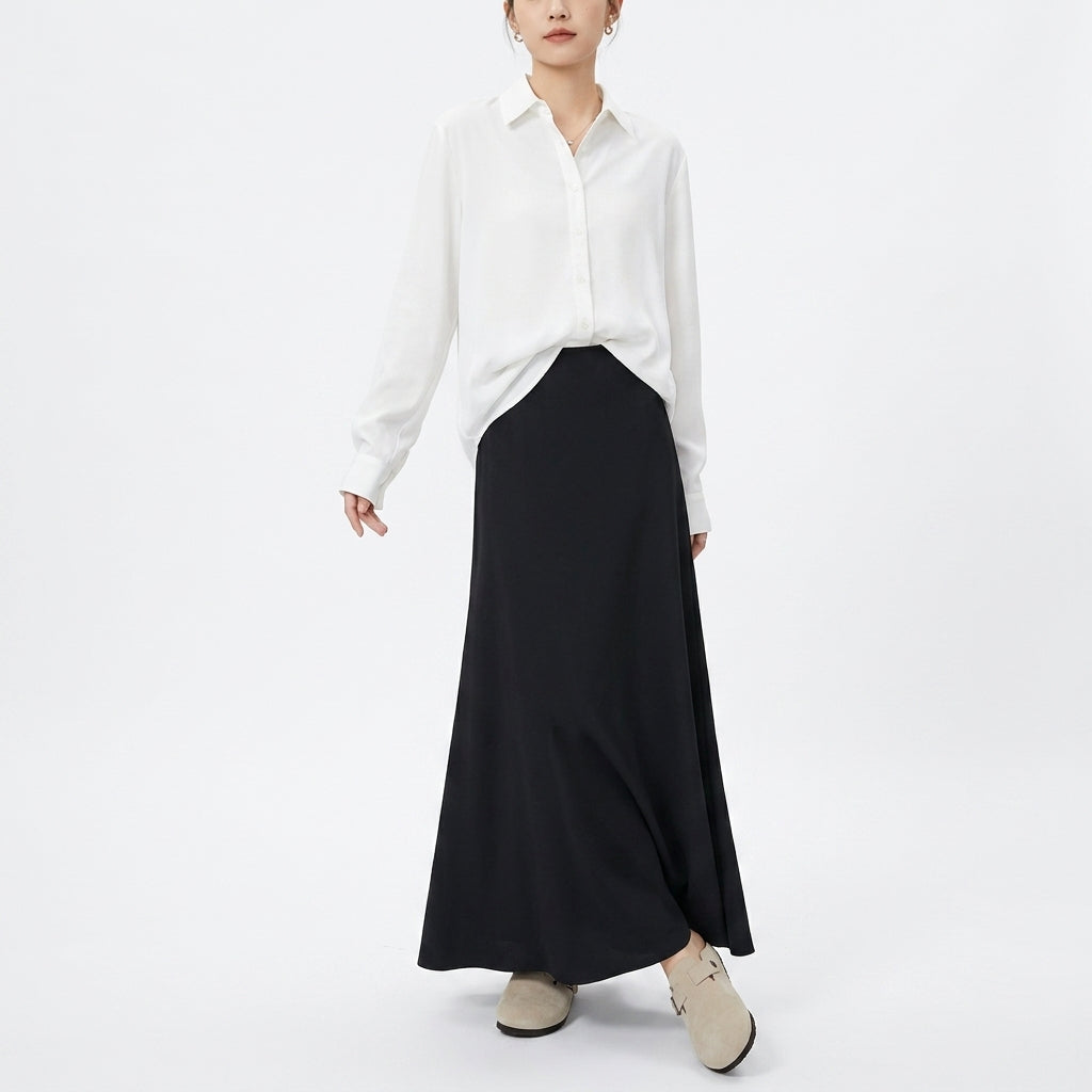 A model wearing a white long-sleeve button-up shirt and a long black skirt, paired with beige loafers, against a plain white studio background.