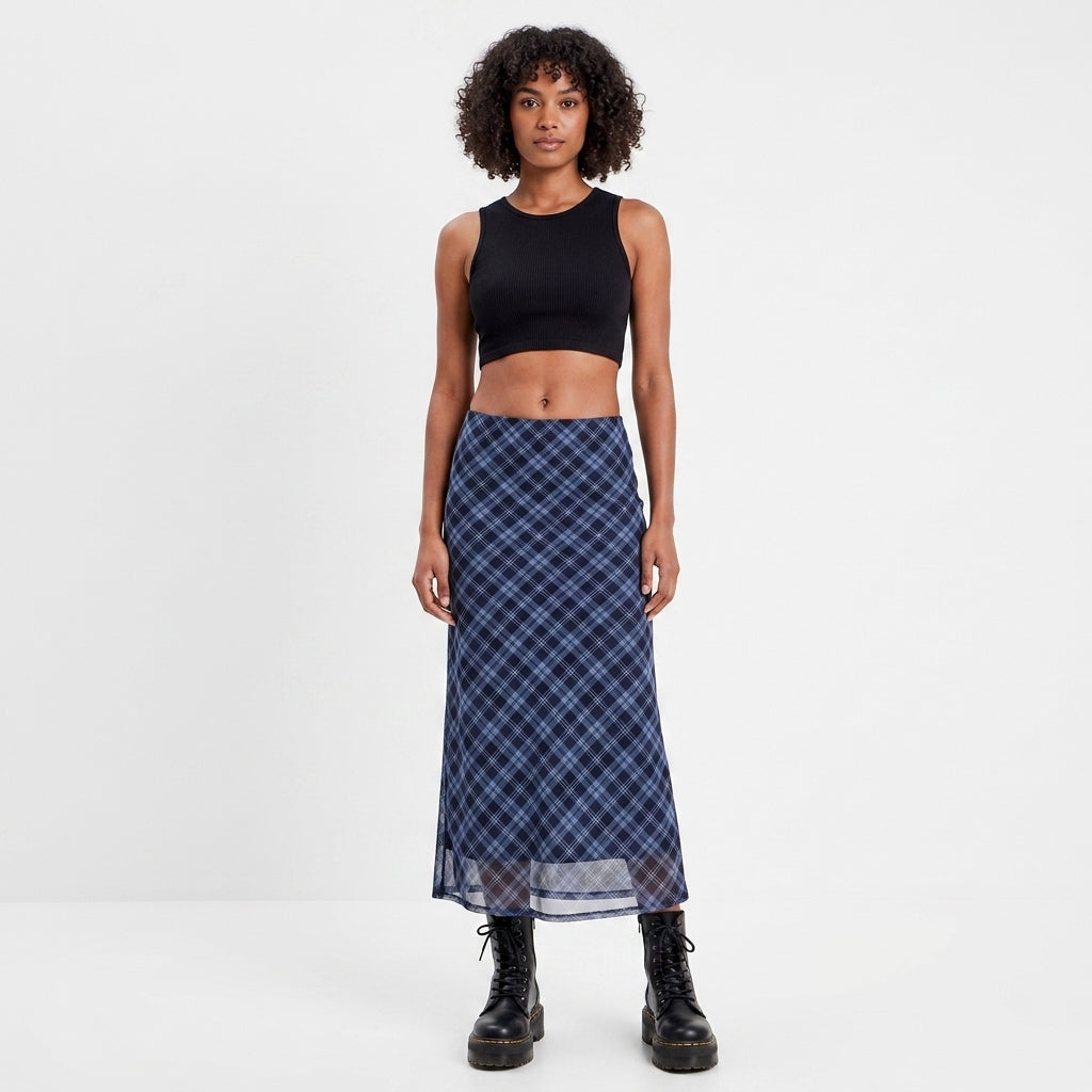 A model stands against a plain white studio background wearing a sleeveless black crop top and a long navy plaid skirt with black combat boots.