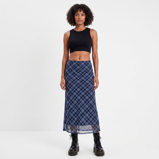 A model stands against a plain white studio background wearing a sleeveless black crop top and a long navy plaid skirt with black combat boots.