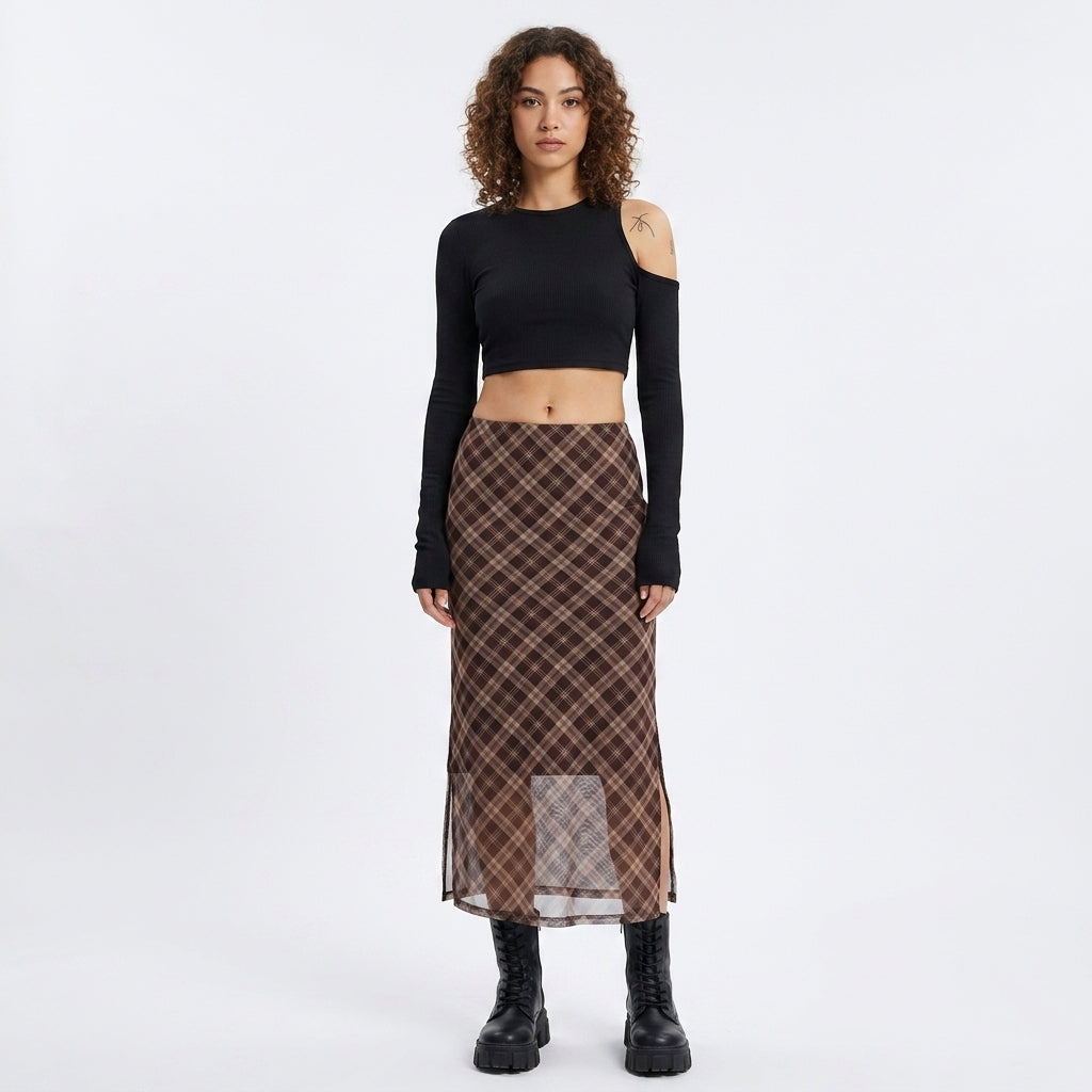 A model stands against a light gray studio background wearing a black long-sleeve crop top with cutout shoulders and a brown plaid midi skirt with a sheer lower panel, paired with black boots.
