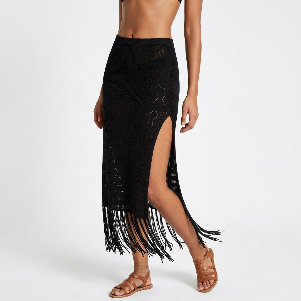 A black crochet knit midi skirt with fringe and a high side slit, shown on a model wearing brown strappy sandals against a light gray studio background.