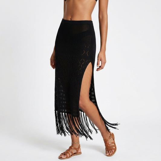 A black crochet knit midi skirt with fringe and a high side slit, shown on a model wearing brown strappy sandals against a light gray studio background.
