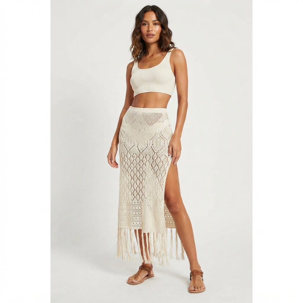 A woman modeling a two-piece cream knit set consisting of a sleeveless crop top and a high-waisted, fringed crochet maxi skirt with a side slit, paired with tan flat sandals against a light gray studio background.