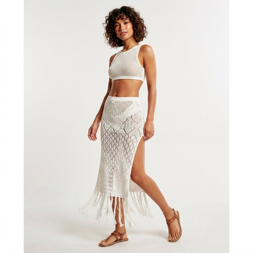 A woman modeling a white crochet two-piece set consisting of a sleeveless crop top and a high-slit midi skirt with fringe, paired with tan strappy sandals against a plain light background.