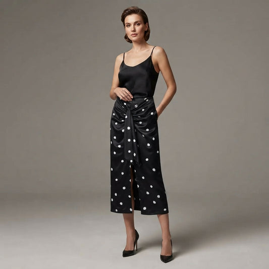 A woman models a black midi wrap skirt with white polka dots and a front slit, paired with a black satin camisole, against a neutral gray studio background.