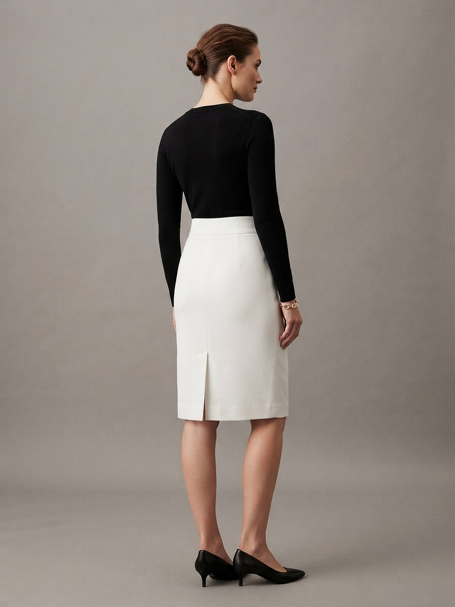 A woman shown from the back wearing a black long-sleeve knit top and a white knee-length skirt with a front slit, paired with black heels, against a plain gray studio background.