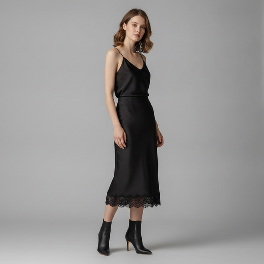 A woman modeling a black satin midi slip dress with thin straps and a lace hem, paired with black ankle boots, against a solid gray studio background.